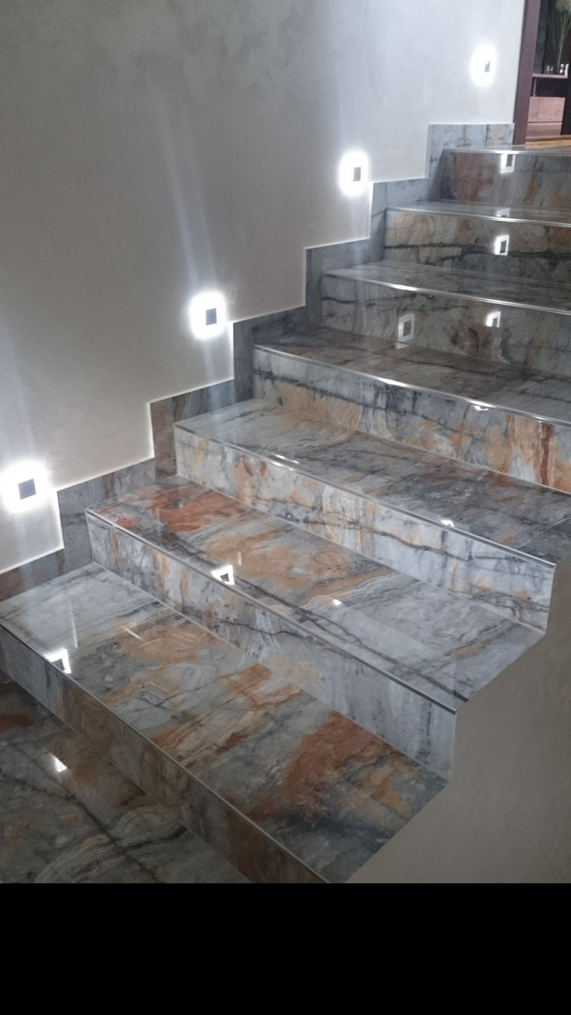 Wykończone schody z płytek imitujących marmur, oświetlone kwadratowymi lampami ściennymi, z widocznymi detalami wykończenia krawędzi.