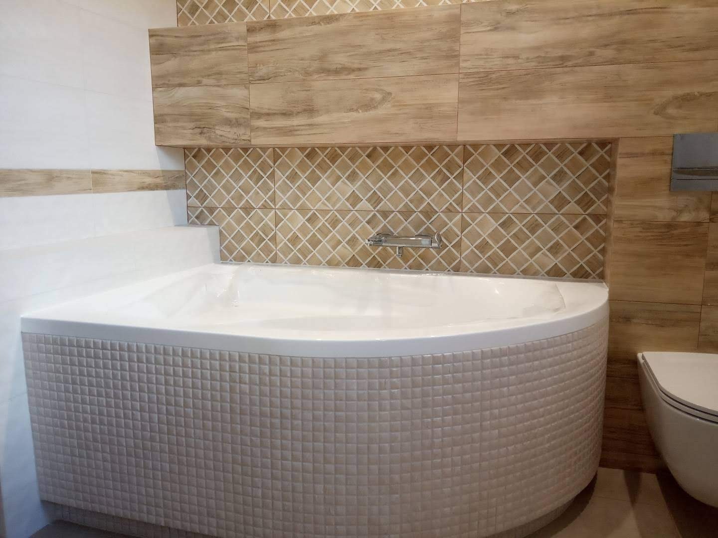 Nowoczesna łazienka z narożną wanną obudowaną mozaiką, drewnopodobne płytki na ścianie, armatura chromowana.