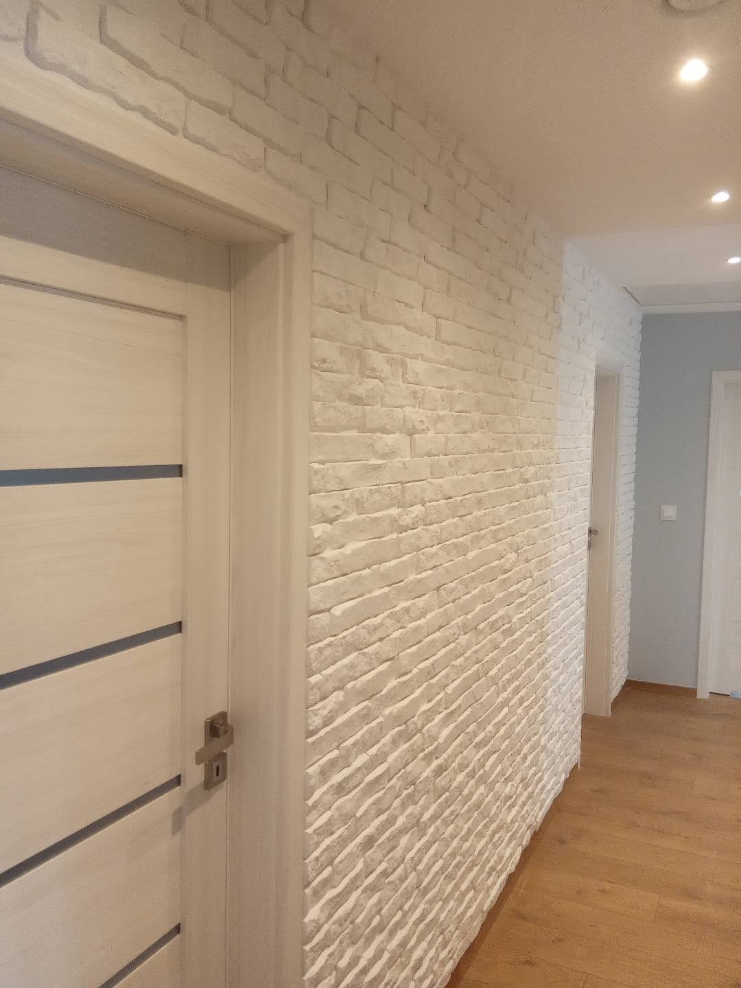 Ściana wykończona dekoracyjną białą cegłą z widoczną fakturą, sąsiadująca z białymi drzwiami z poziomymi, ciemnymi wstawkami, w tle fragment korytarza z drewnianą podłogą i oświetleniem punktowym.