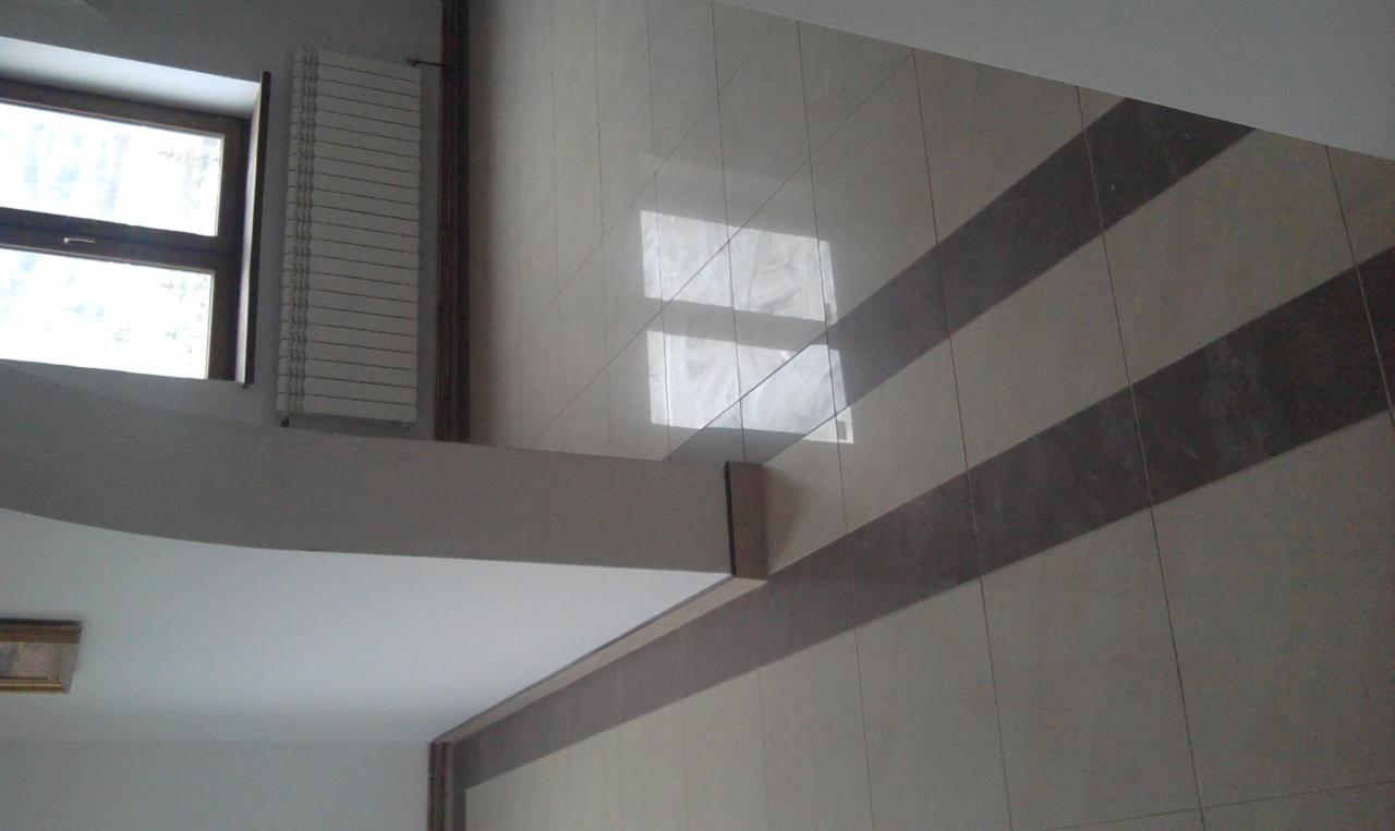 Ściana wyłożona jasnymi płytkami ceramicznymi z poziomymi pasami ciemniejszych płytek, widoczny fragment okna i grzejnika.