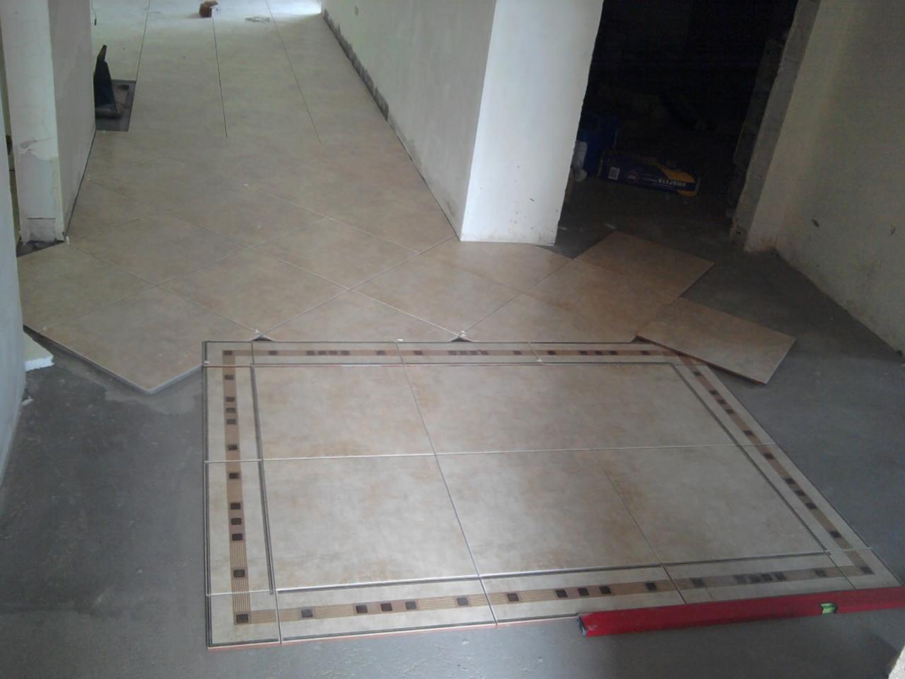 Układanie beżowych płytek podłogowych z dekoracyjnym obramowaniem, poziomica w prawym dolnym rogu, widoczne krzyżyki dystansowe.