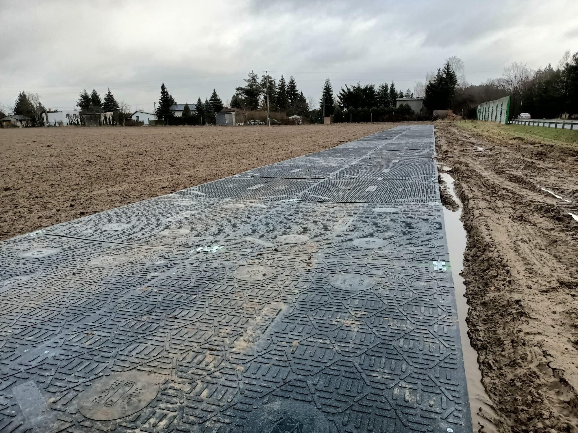 Ułożone, czarne płyty na błotnistym terenie, tworzące tymczasową drogę. Widoczne ślady opon i kałuże. W tle drzewa i szare niebo.