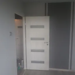 Wnętrze pokoju z szarą ścianą z ciemnymi pasami, drewnianą podłogą i białymi drzwiami z przeszkleniami; w tle widoczne środki czystości.