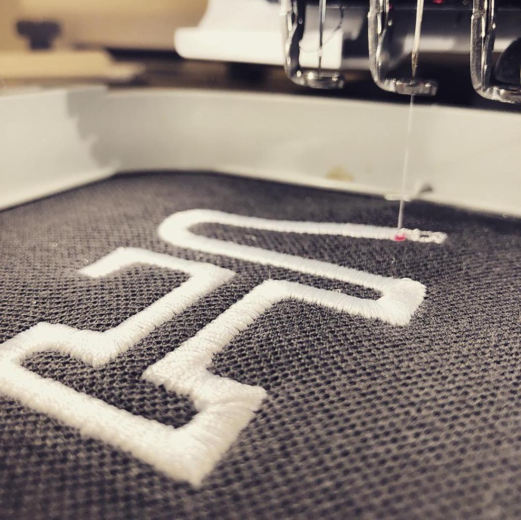 Proces tworzenia białego, geometrycznego wzoru na ciemnoszarym materiale za pomocą maszyny do szycia, z widoczną igłą i nitką.