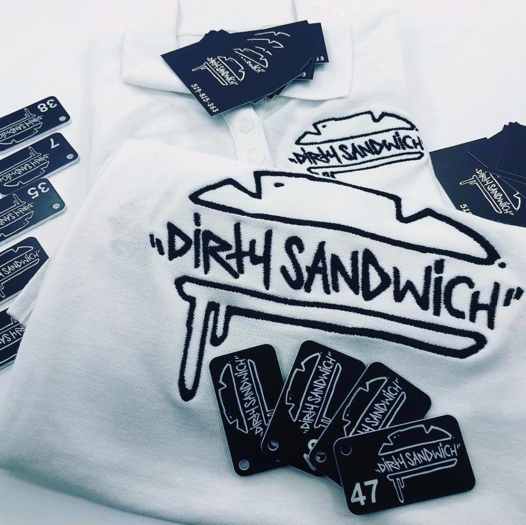 Biała koszulka polo z wyhaftowanym czarnym logo 'Dirty Sandwich' na piersi i kołnierzyku, otoczona czarnymi kartami z tym samym logo i numerami.