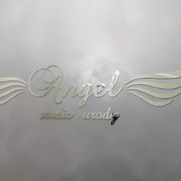 Metalowy napis 'Angel studio urody' z dekoracyjnymi skrzydłami umieszczony na szarej, teksturowanej ścianie.