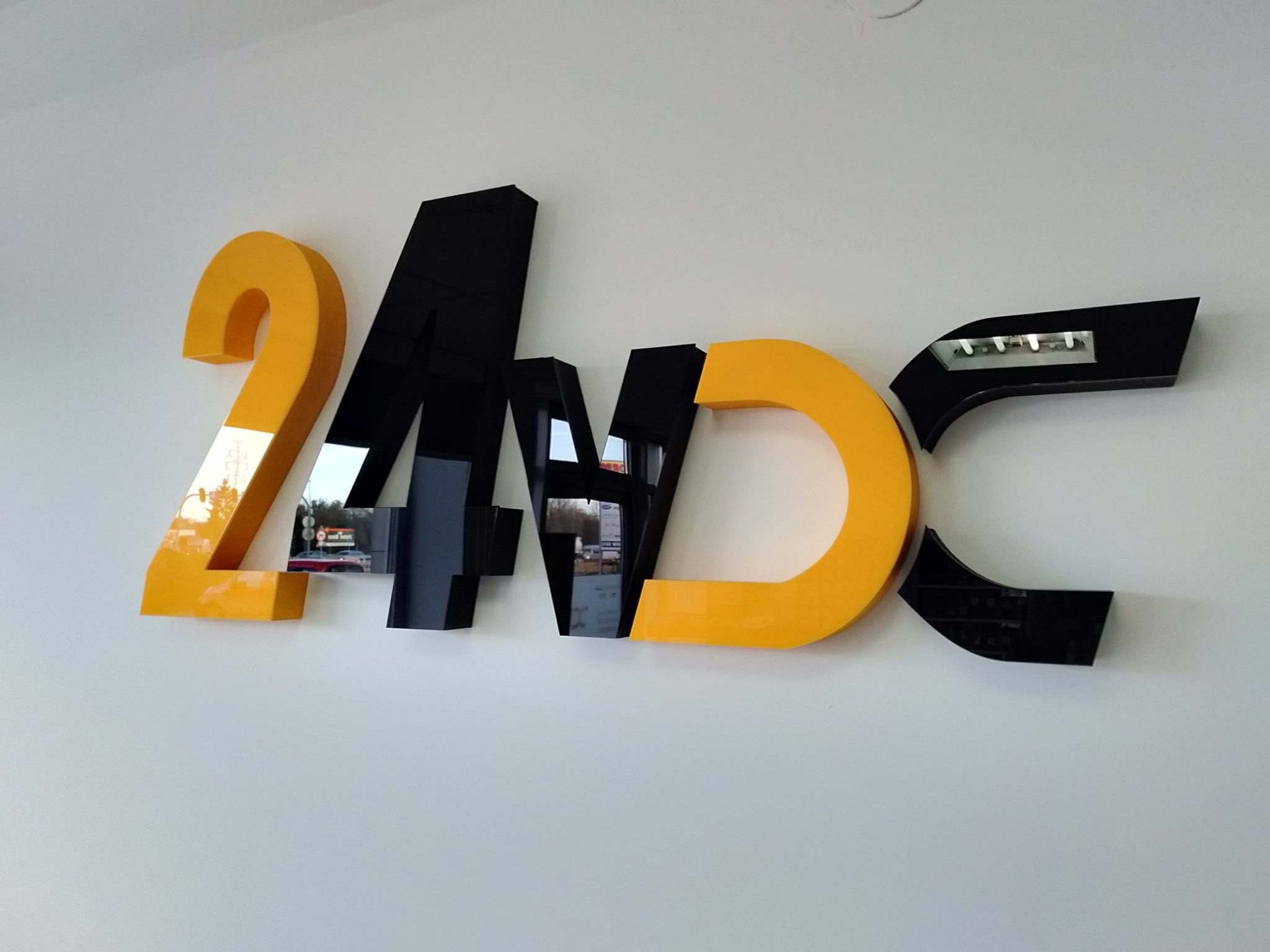 Przestrzenne litery 24/7DC w kolorach żółtym i czarnym, zamontowane na białej ścianie. Litery z efektem lustrzanym, odbijają otoczenie.