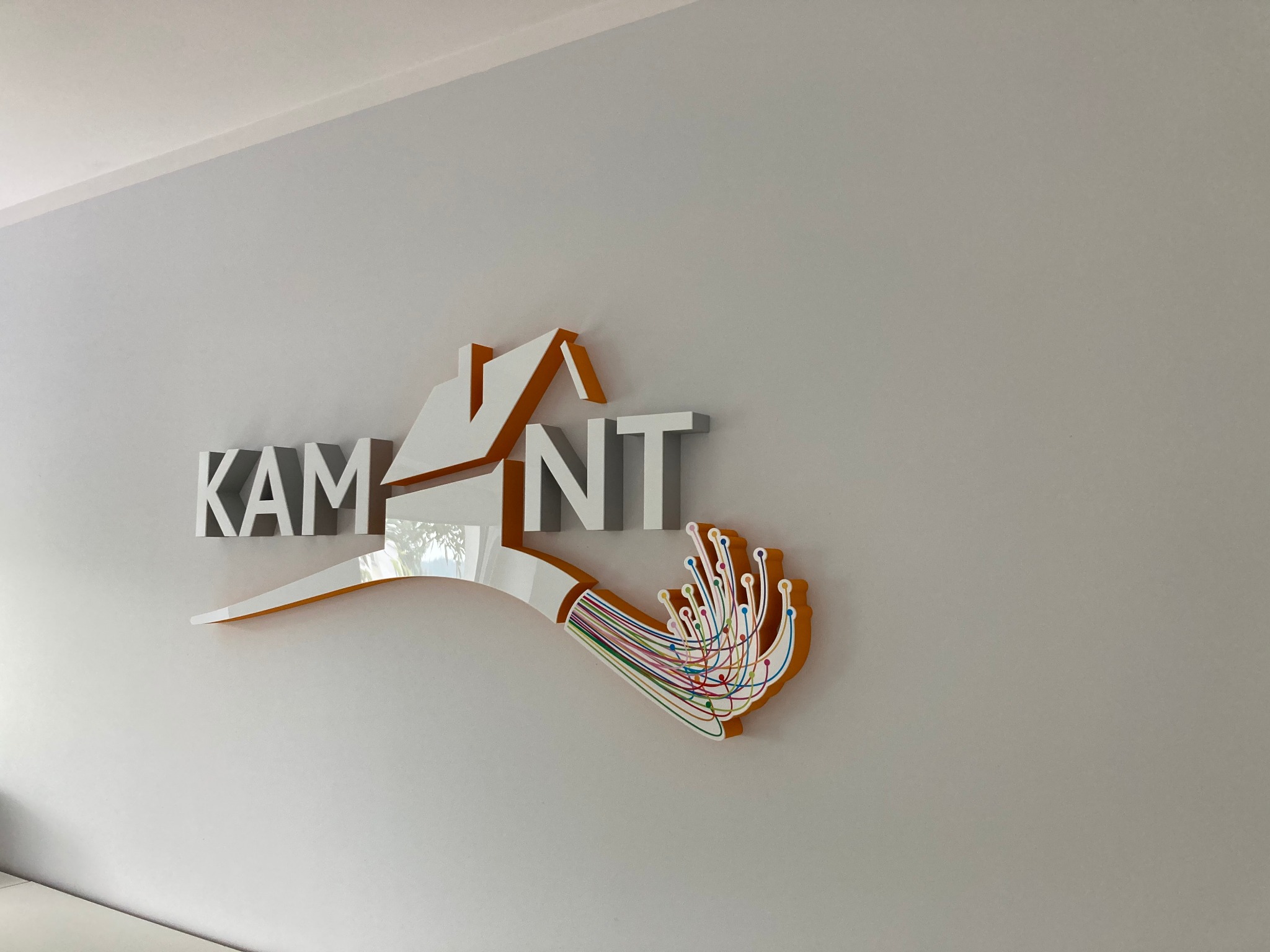 Logo firmy 'KAM NT' wykonane z tworzywa sztucznego, z motywem wiązki kabli światłowodowych w kolorach tęczy, zamontowane na jasnoszarej ścianie.