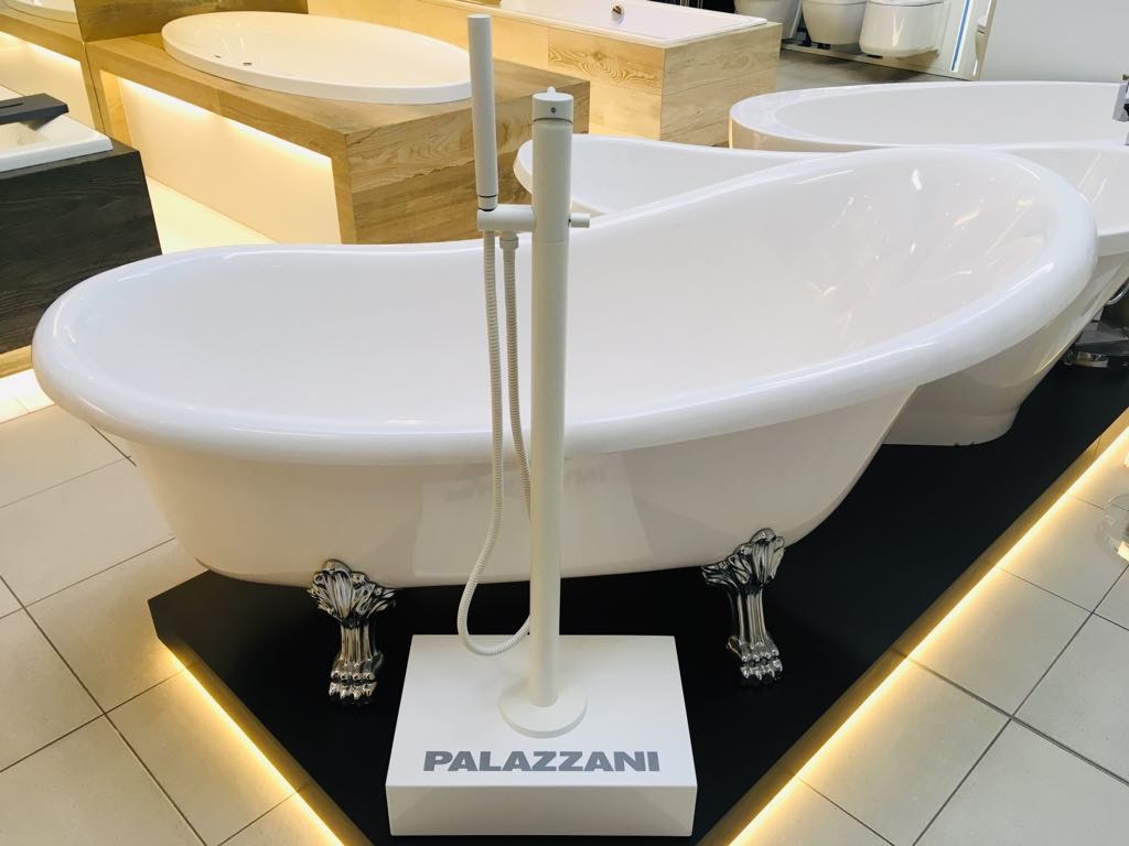 Wolnostojąca, biała wanna na ozdobnych, srebrnych nóżkach w kształcie lwich łap, z białą baterią podłogową i słuchawką prysznicową, prezentowana na czarnym podeście z logo 'PALAZZANI'.