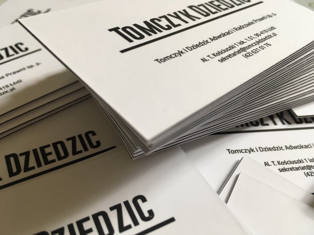 Stos wizytówek z czarnym, pogrubionym tekstem 'Tomczyk Dziedzic' i danymi kontaktowymi kancelarii adwokackiej, ułożonych pod kątem, widoczne krawędzie papieru.