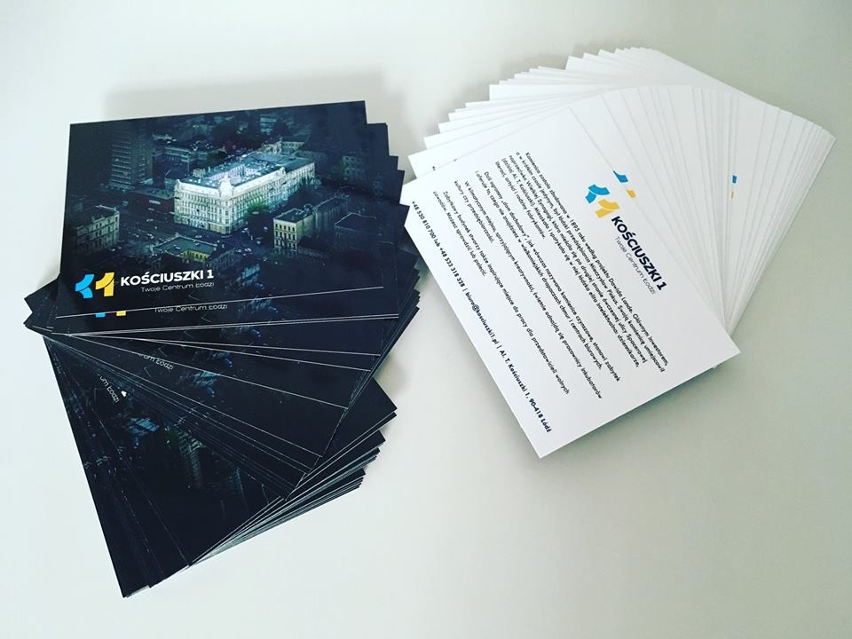 Stos ulotek i wizytówek z logo Kościuszki 1, prezentujących budynek na nocnym zdjęciu lotniczym, ułożonych na białym tle.