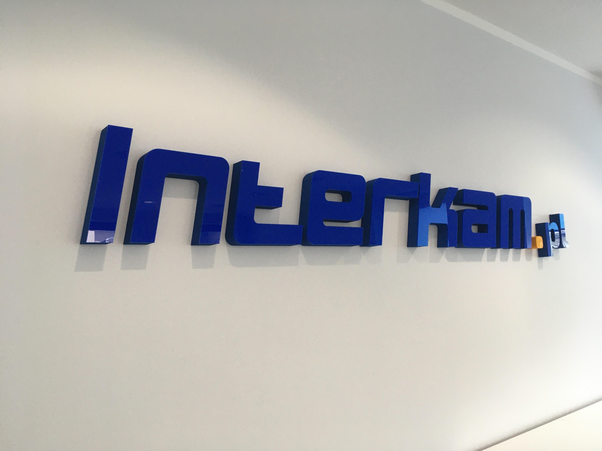 Niebieski, przestrzenny napis firmowy 'Interkan' zamontowany na białej ścianie, z widocznym fragmentem oświetlenia w tle.