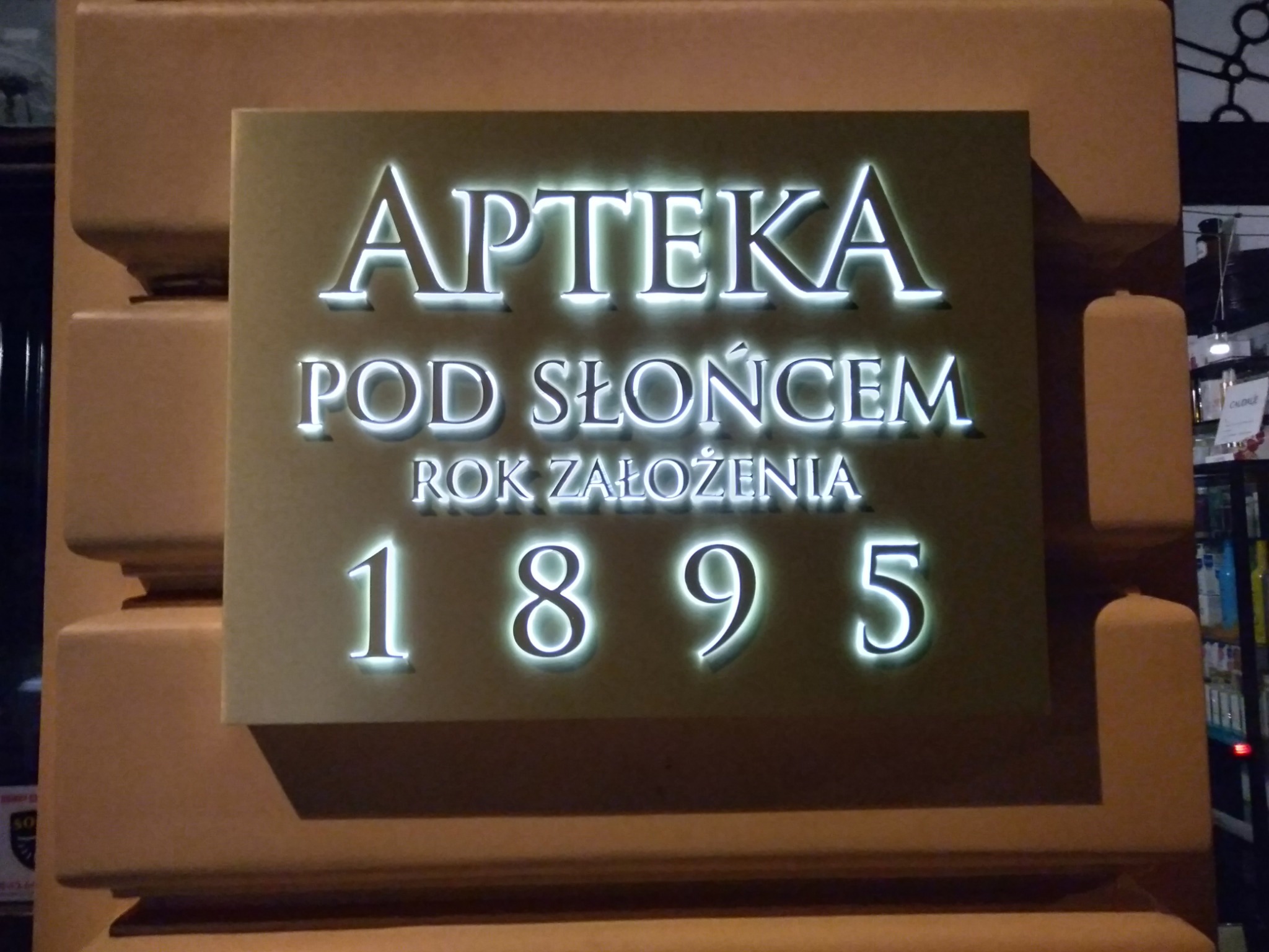 Podświetlany szyld apteki 'Apteka Pod Słońcem', z datą założenia 1895, zamontowany na brązowym filarze, widoczny fragment wnętrza apteki w tle.