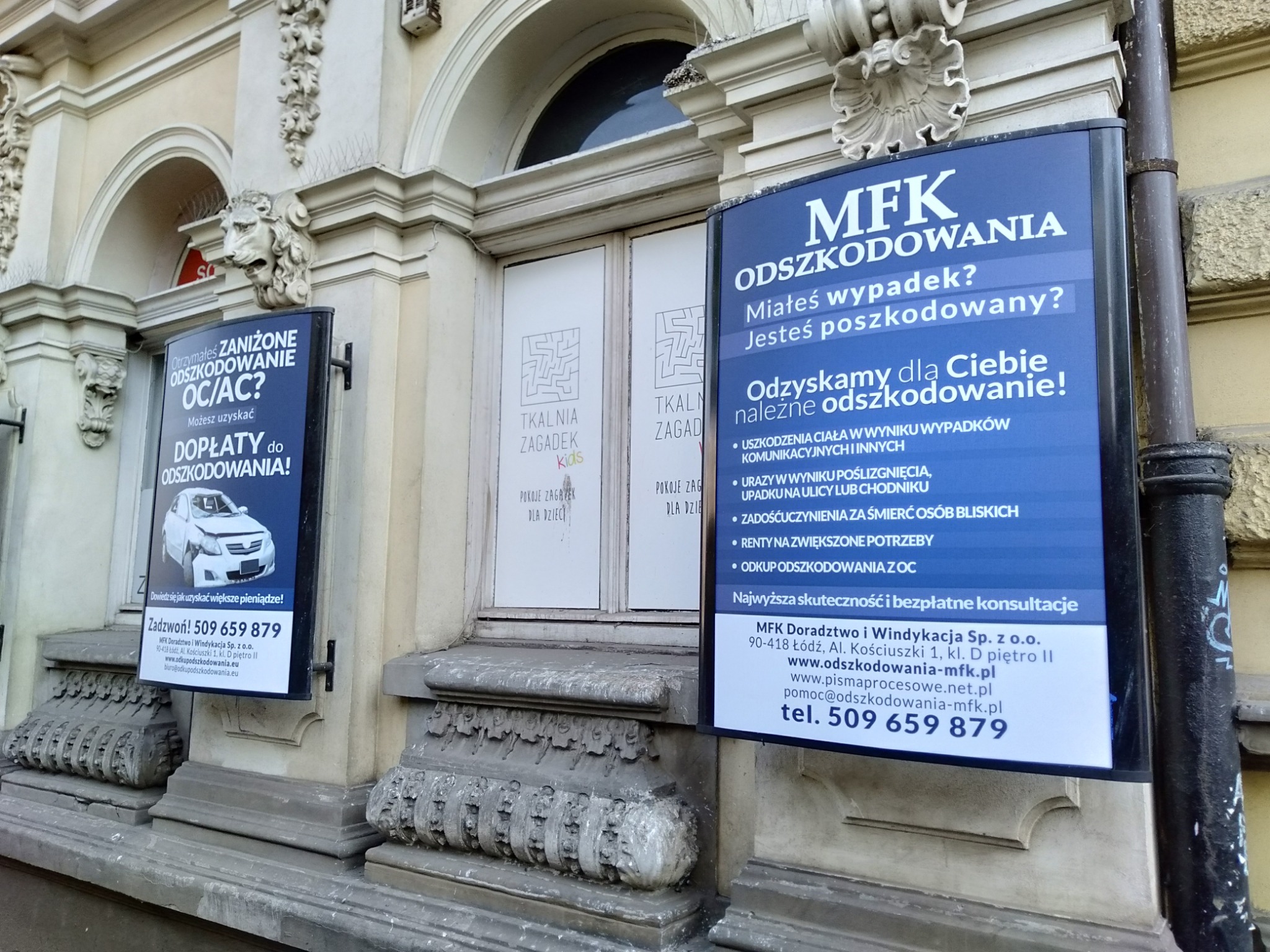 Dwa plakaty reklamowe firmy MFK Doradztwo i Windykacja, oferującej pomoc w uzyskaniu odszkodowań, umieszczone na elewacji kamienicy z dekoracyjnymi elementami architektonicznymi. Jeden plakat...