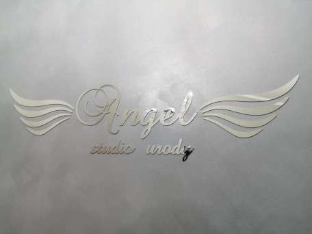 Metalowy napis 'Angel studio urody' z dekoracyjnymi skrzydłami umieszczony na szarej, teksturowanej ścianie.