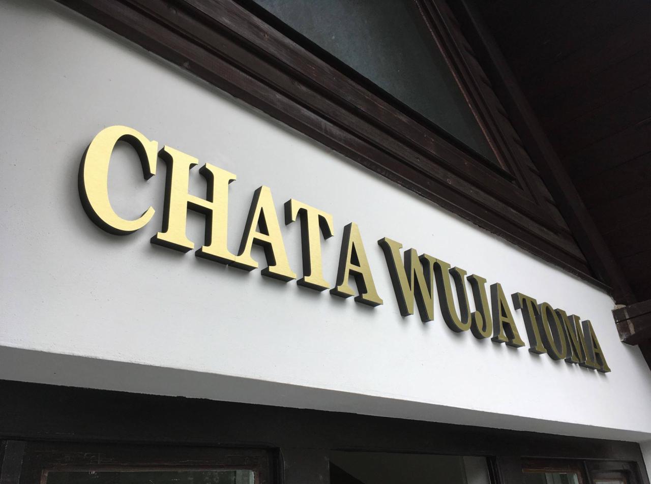 Złote, przestrzenne litery z czarnym cieniem tworzące napis 'CHATA WUJA TOMA' zamontowane nad wejściem do budynku z drewnianą konstrukcją dachu.