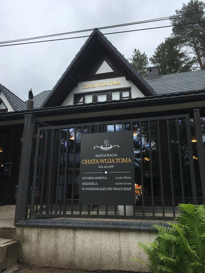 Czarny szyld informacyjny z nazwą restauracji 'Chata Wuja Toma' i godzinami otwarcia, umieszczony przed budynkiem o charakterystycznej architekturze góralskiej z ciemnym dachem i jasną fasadą...