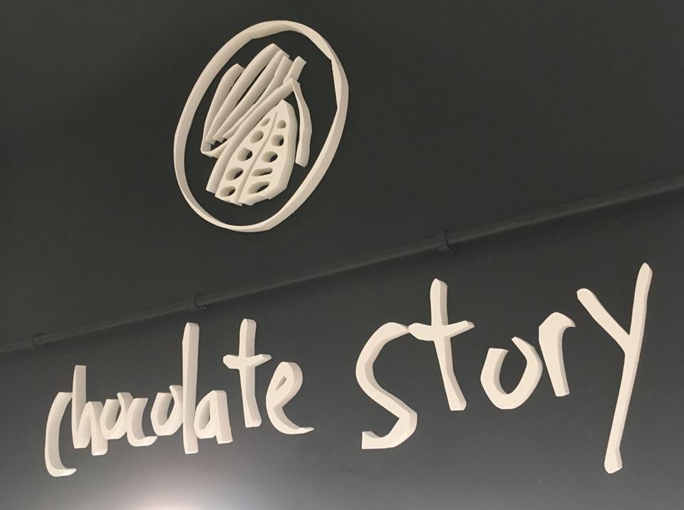 Białe, trójwymiarowe logo przedstawiające ziarno kakaowca w okręgu oraz napis 'chocolate story' umieszczone na ciemnoszarej ścianie.
