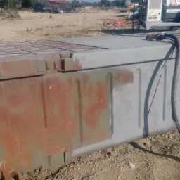 Fragment dużej metalowej konstrukcji, częściowo oczyszczonej z rdzy, z widocznym wężem do czyszczenia, na tle ziemi i pojazdów.