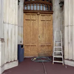 Drewniane, dwuskrzydłowe drzwi zabytkowe z dekoracyjnym nadświetlem w Legnicy, przed którymi leży zwinięty wąż i stoi drabina, sugerujące prace renowacyjne.