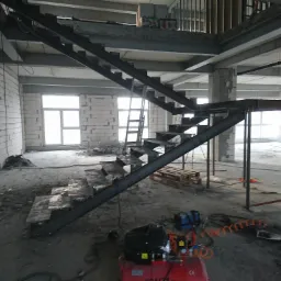 Surowe metalowe schody w trakcie montażu w surowym wnętrzu hali, widoczny kompresor i narzędzia na podłodze, ściany z białych bloczków, duże okna w tle.