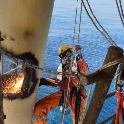 Pracownik w kombinezonie i kasku ochronnym, z maską na twarzy, wykonuje prace na wysokości na platformie wiertniczej, iskry lecą podczas obróbki metalu, w tle widoczne spokojne morze.