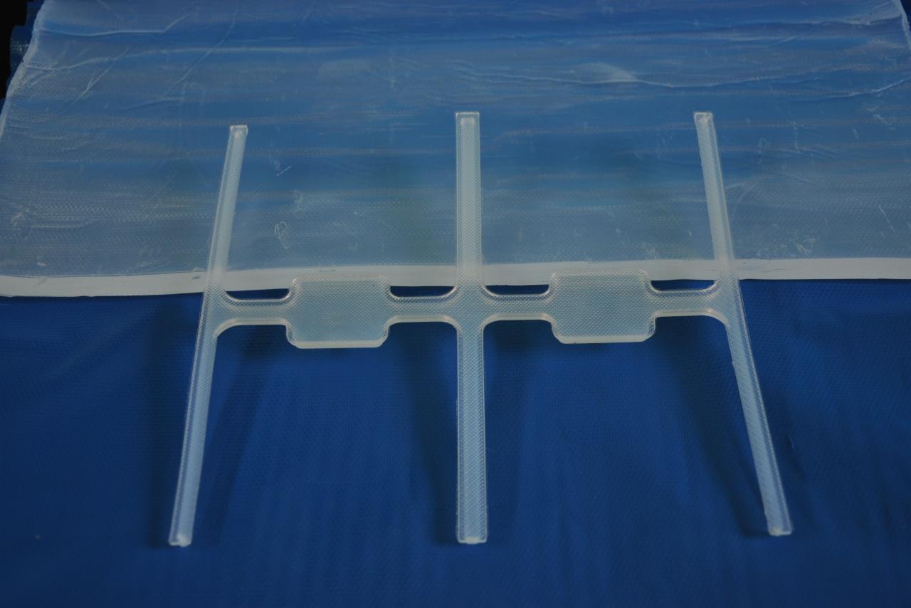 Przezroczysty plastikowy element montażowy z czterema pionowymi prętami na niebieskim tle, prawdopodobnie do mocowania folii lub innych materiałów.