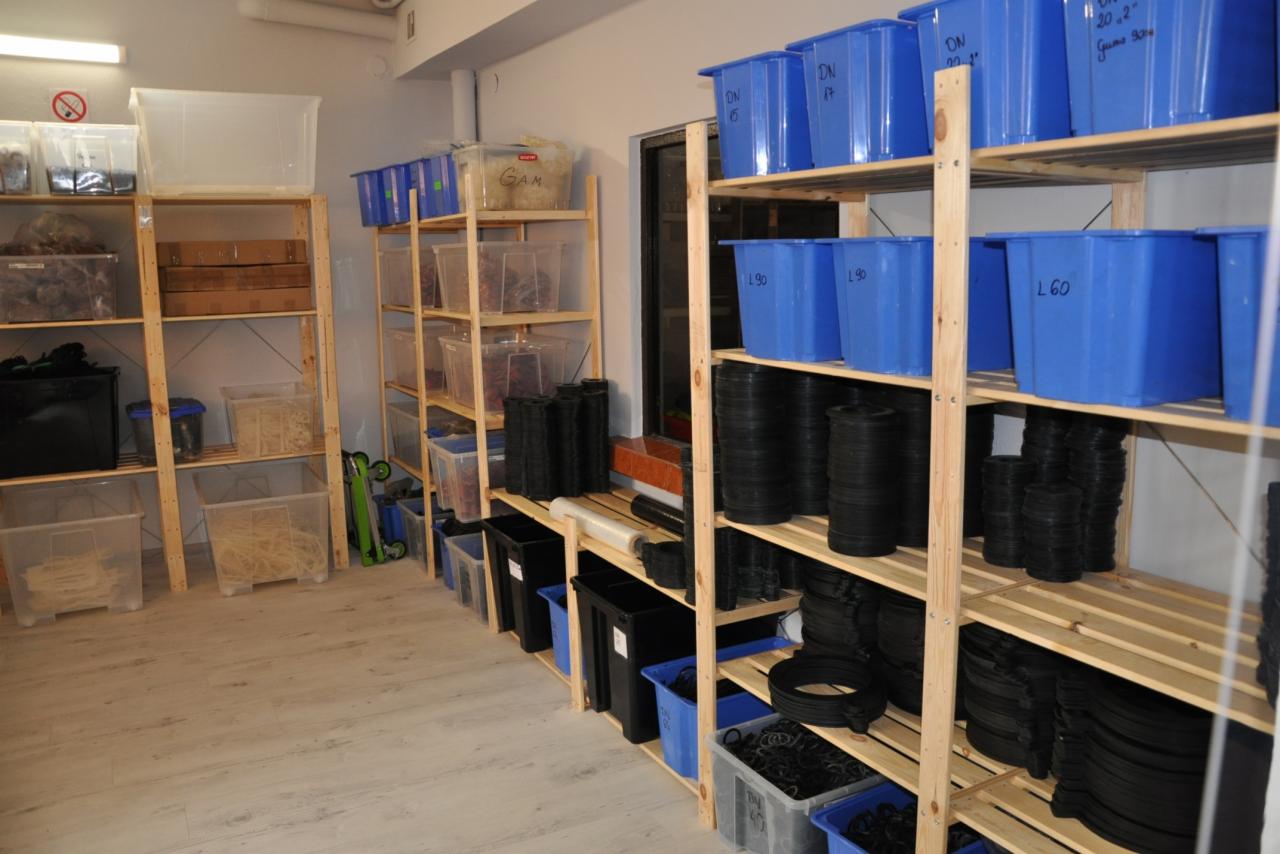 Magazyn z drewnianymi regałami wypełnionymi przezroczystymi i niebieskimi pojemnikami z różnymi materiałami instalacyjnymi, w tym czarnymi uszczelkami i rurami.
