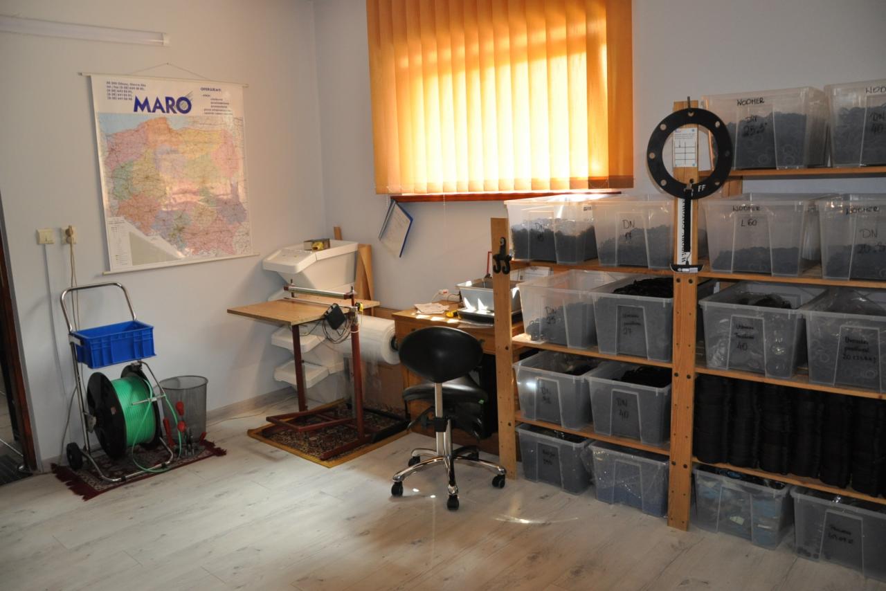 Wnętrze magazynu lub sklepu z armaturą: regał z przezroczystymi pojemnikami zawierającymi czarne uszczelki i złączki, w tle biurko i plakat z mapą.