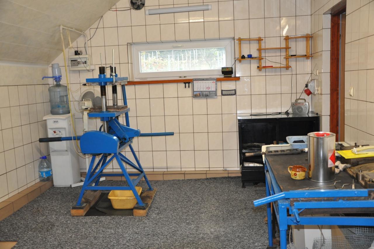 Warsztatowe wnętrze z niebieską prasą hydrauliczną, dystrybutorem wody i stołem roboczym z narzędziami, na tle wykafelkowanych ścian.