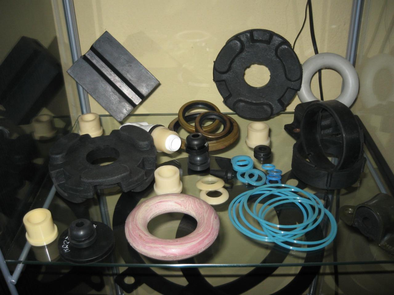 Różnorodne gumowe i plastikowe uszczelki, pierścienie, tuleje i podkładki w odcieniach czerni, bieli, brązu i błękitu, ułożone na szklanej półce w gablotce.