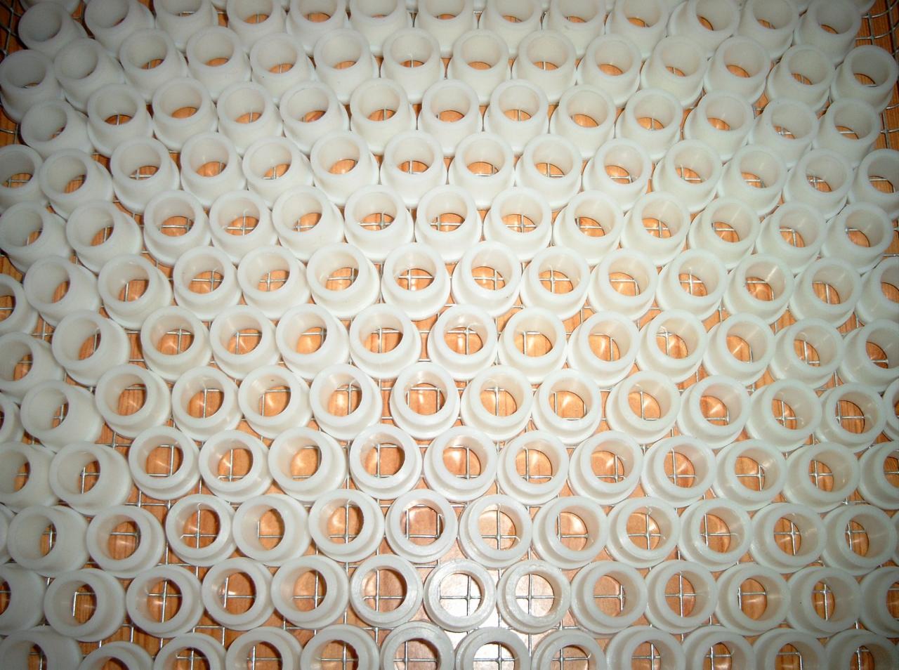 Równomiernie ułożone białe, plastikowe tuleje na metalowej siatce, widoczne delikatne cienie, podkreślające trójwymiarowość elementów.