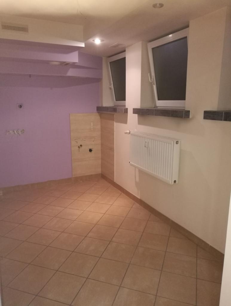 Pusty pokój z fioletową ścianą, brązowymi płytkami podłogowymi, dwoma oknami z białymi ramami i grzejnikiem na ścianie.