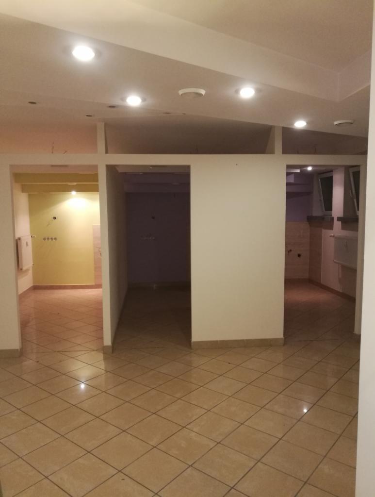 Puste wnętrze mieszkania z beżową podłogą w kratkę i białymi ścianami, widoczne przejścia do innych pomieszczeń, sufit z punktowym oświetleniem.