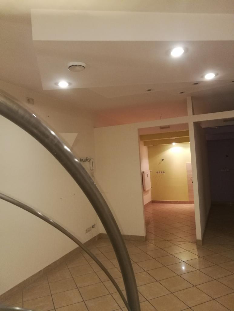 Wnętrze pustego mieszkania z widoczną metalową balustradą schodów, brązową podłogą z płytek i oświetleniem sufitowym.