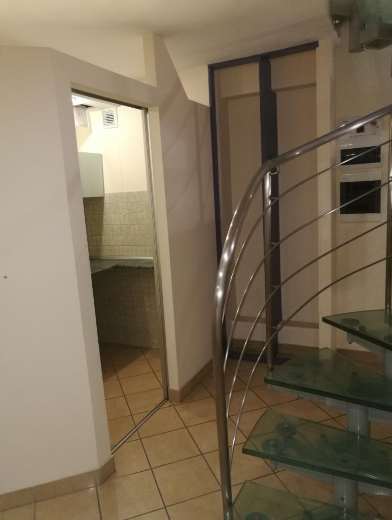 Wnętrze mieszkania z widoczną kuchnią przez otwarte drzwi i szklanymi schodami spiralnymi z metalową poręczą.