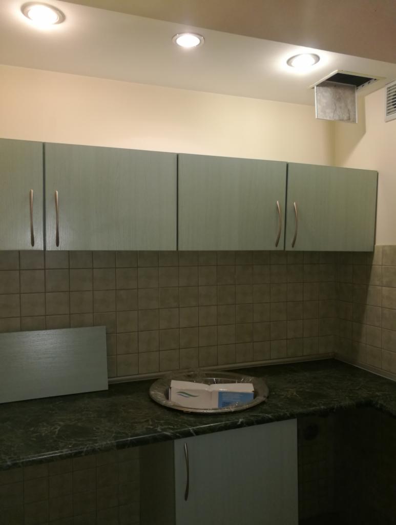 Kuchnia z zielonymi szafkami, zielonym blatem i beżowymi płytkami, na blacie srebrna patera z opakowaniami, widoczne trzy lampy w suficie i kratka wentylacyjna.