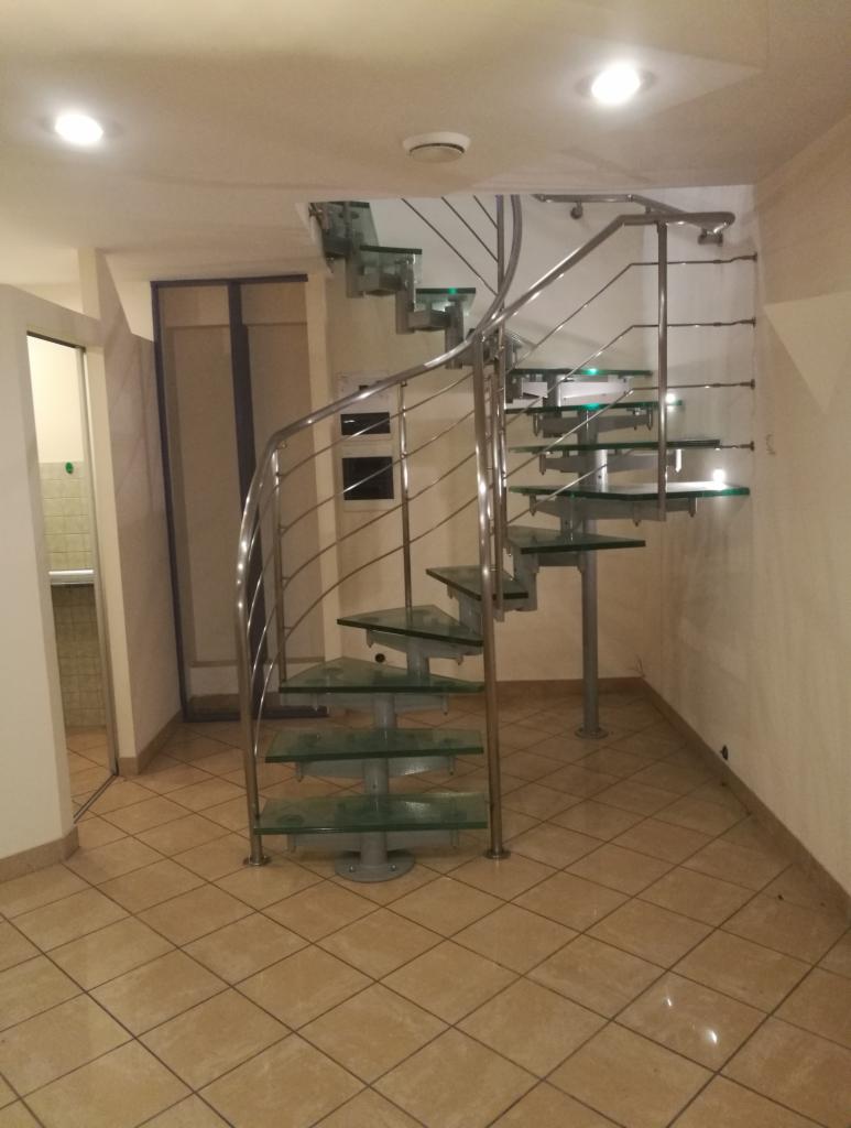 Szklane schody spiralne z metalową balustradą w jasnym wnętrzu z beżową podłogą z płytek.