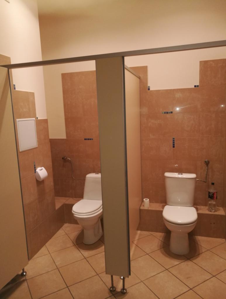 Publiczna toaleta z dwoma kabinami, płytki ceramiczne w odcieniach brązu, widoczna butelka po napoju obok jednej z toalet.