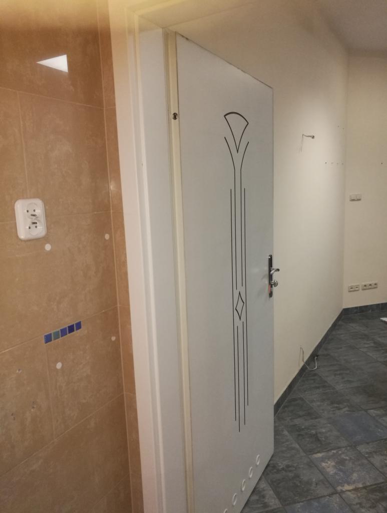 Białe drzwi wewnętrzne z dekoracyjnym wzorem w kształcie rombu i stylizowanych liści na tle wykończonej łazienki z brązowymi płytkami i niebieskim akcentem oraz fragmentu korytarza z szarą podłogą.