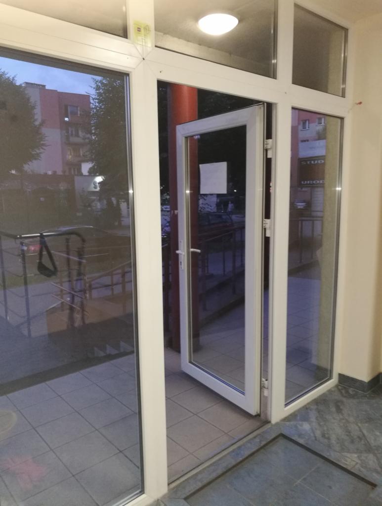 Otwarta, biała, przeszklona witryna wejściowa do budynku usługowego z widokiem na chodnik i budynki w tle.