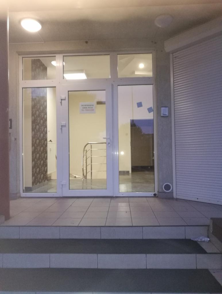 Wejście do biura z trzema schodami, białe drzwi z napisem 'Do wynajęcia lokal piwn.', roleta zewnętrzna częściowo opuszczona.