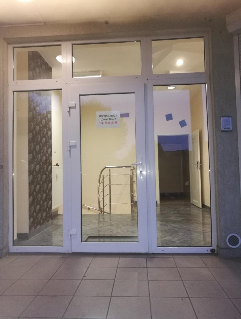 Wejście do lokalu z białymi drzwiami i ramami, na drzwiach kartka 'Do wynajęcia', wewnątrz widoczna klatka schodowa z metalową barierką.