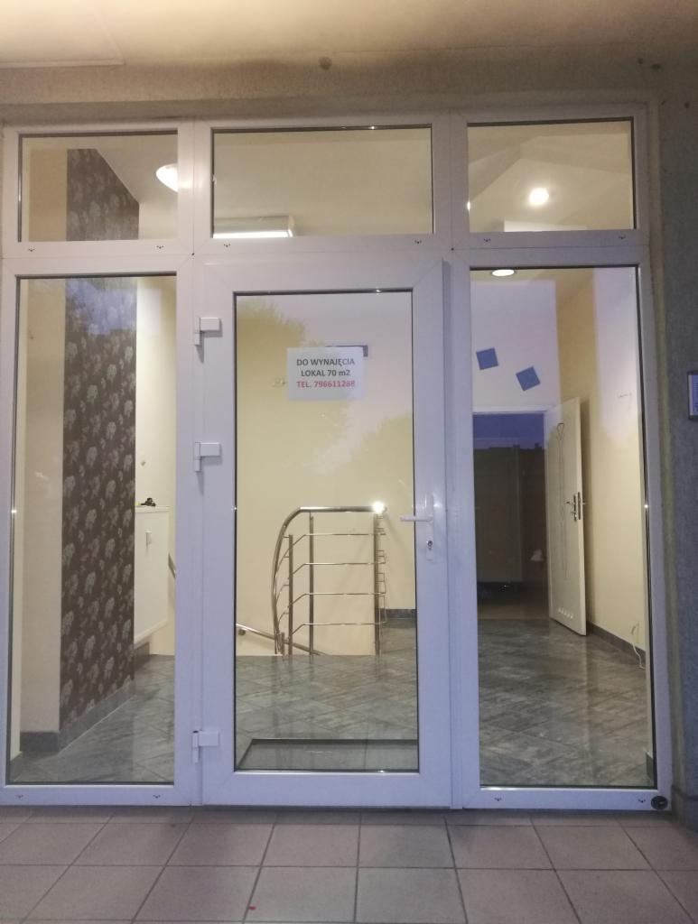 Wejście do lokalu z informacją o wynajmie, widoczna klatka schodowa z metalową balustradą i płytki na podłodze.