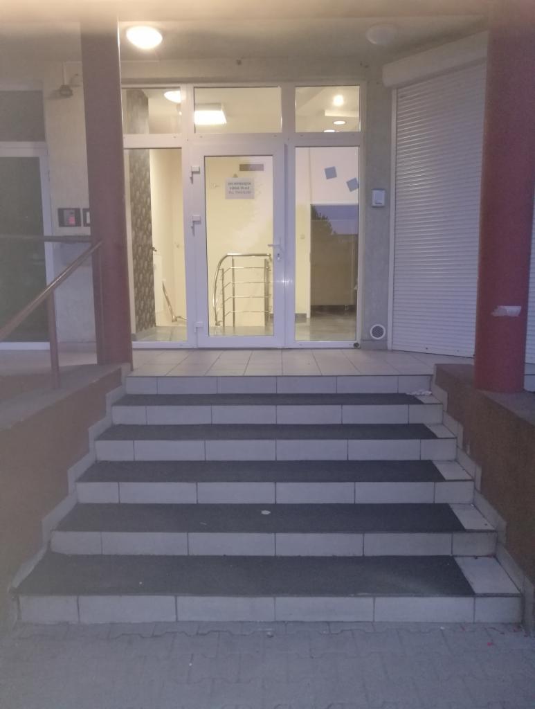 Wejście do budynku z białymi drzwiami i schodami wyłożonymi szarymi płytkami, widoczne oświetlenie sufitowe i częściowo zasłonięte rolety.
