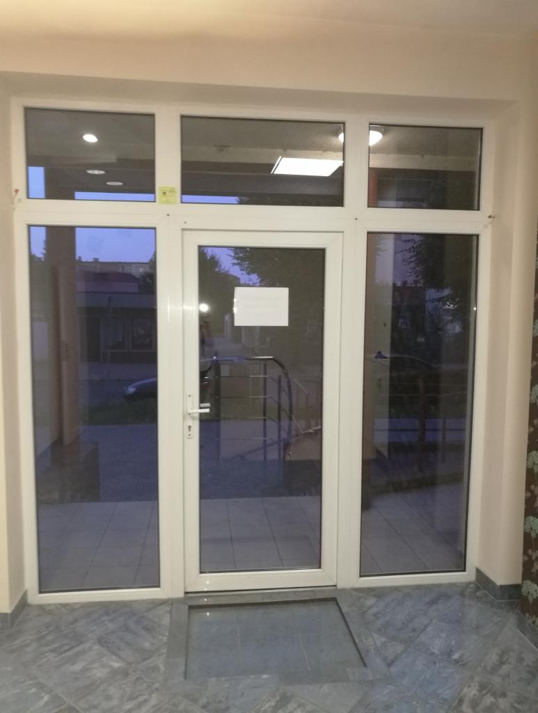 Białe aluminiowe drzwi wejściowe z szybami, widok z wnętrza budynku na zewnątrz, szare płytki podłogowe.