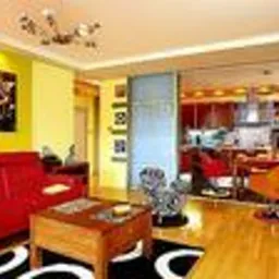 Jasne wnętrze po remoncie: żółte ściany, czerwona sofa, drewniana podłoga, wzorzysty dywan w czarno-białe okręgi, otwarta przestrzeń na kuchnię z czerwonymi krzesłami barowymi i pomarańczowym...