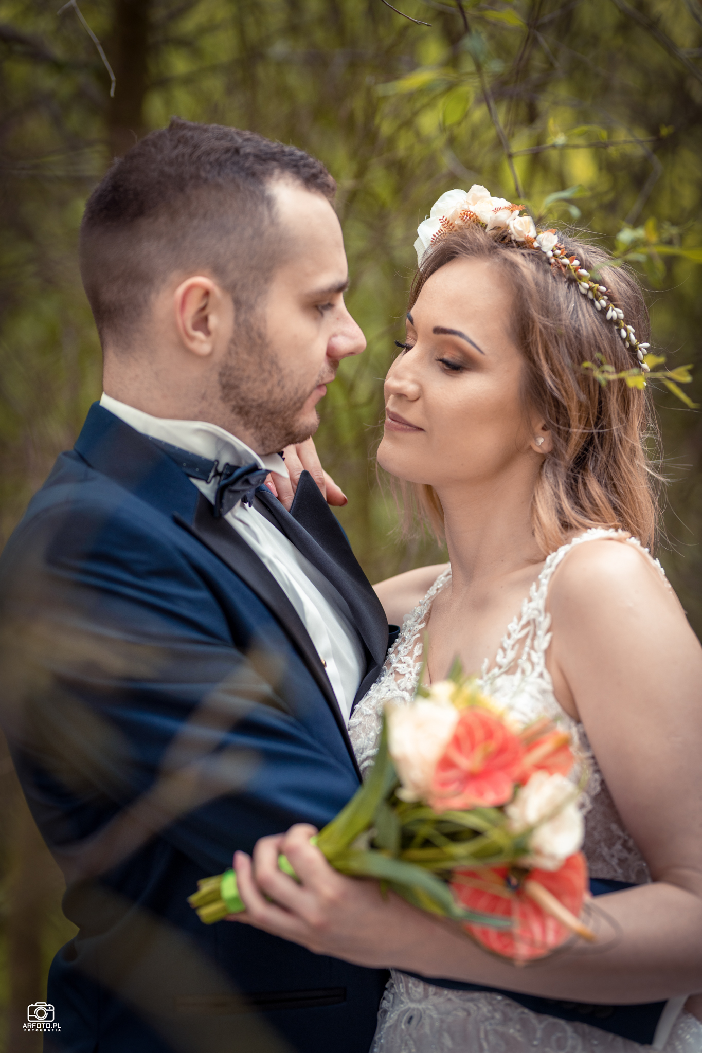 Elegancko ubrana para młoda, panna młoda w wianku z kwiatów i z bukietem w rękach, patrzy na pana młodego w granatowym smokingu, na tle drzew.