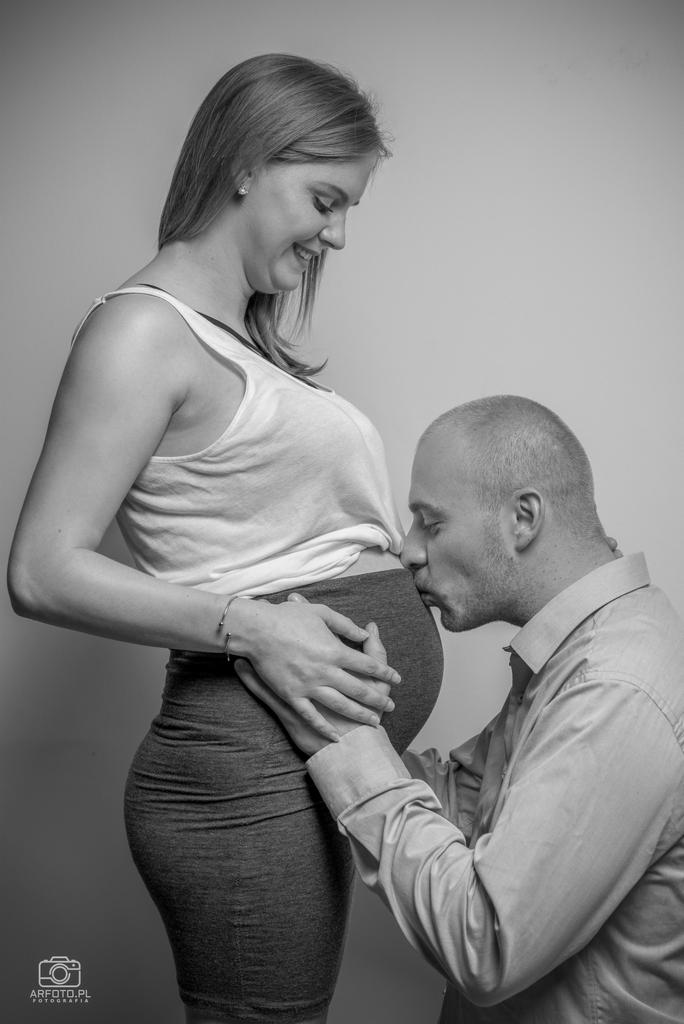 Czarno-białe zdjęcie kobiety w ciąży, partner całuje jej brzuch, oboje uśmiechnięci, ona w białym topie i szarej spódnicy, on w jasnej koszuli, proste tło.