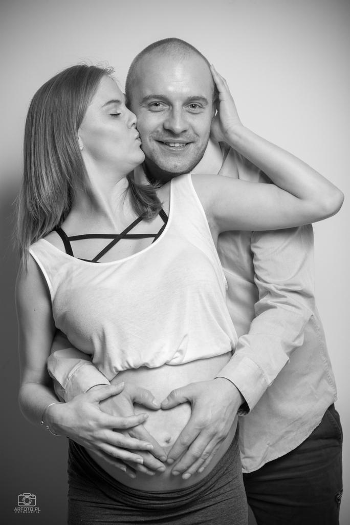 Czarno-białe zdjęcie przyszłej mamy, całowanej w policzek przez partnera, oboje trzymają ręce na brzuchu kobiety, formując kształt serca.